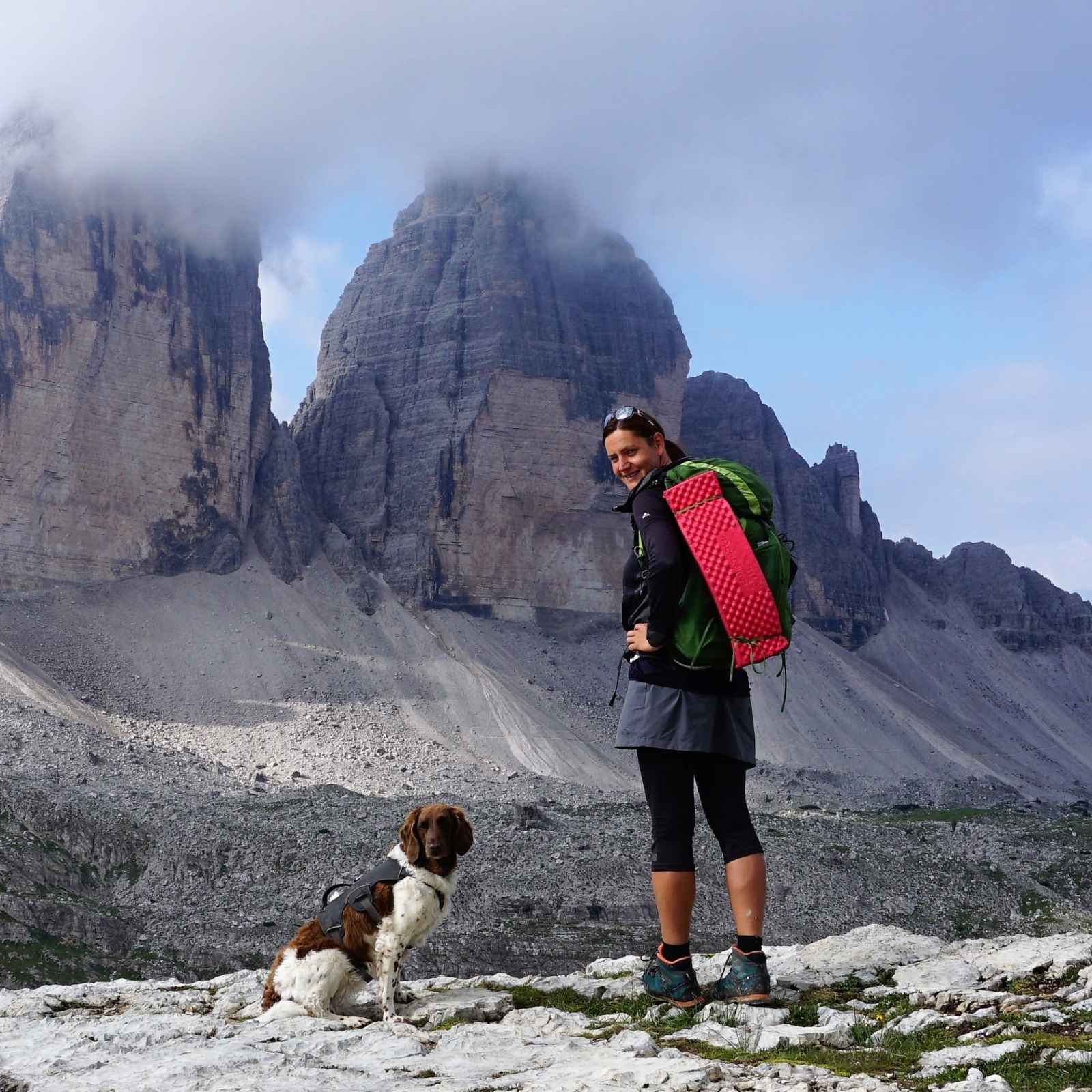 A woman wearing a backpack in front of mountain range with her dog